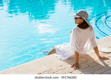 Fashion Beautiful Woman On Summer Vacation Relaxing At Luxury Resort Spa Poolside. Young  Fashionable Lady Wearing Sun Hat And White Kaftan.