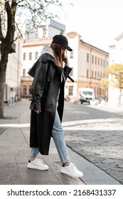 Fashion Beautiful Young Woman Model Hipster In Fashionable Black Casual Clothes With A Cap In A Long Leather Coat With Jeans And White Shoes Walking In The City. Cool Female Urban Outfit Look 