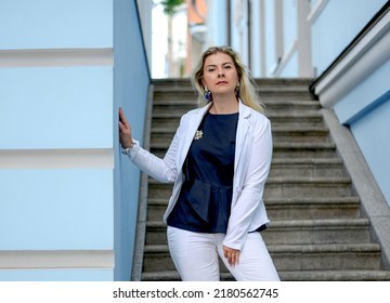 Fashion And Beauty Concept. Confident сaucasian Woman In A White Suit Standing On The Stairs, Looking Into The Camera. Background Of Blue City Buildings.