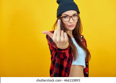 Fashion beauty girl wearing sunglasses, plaid shirt, black beanie hat. Young woman showing middle finger over yellow background.