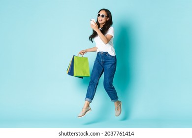 Fashion, Beauty And Lifestyle Concept. Carefree Smiling Beautiful Korean Girl,asian Woman In Sunglasses Looking At Mobile Phone And Holding Shopping Bags While Jumping Over Light Blue Background