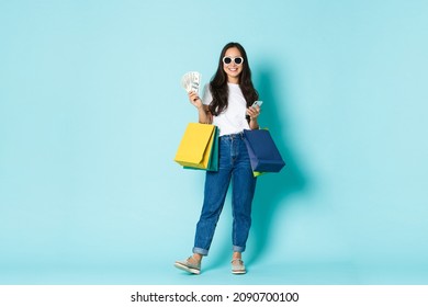 Fashion, Beauty And Lifestyle Concept. Pleased Happy Rich Asian Girl In Sunglasses, Wasting Cash In Mall, Shopping With Huge Sum Of Money, Holding Bags And Mobile Phone, Light Blue Background