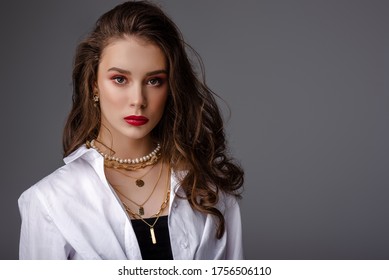 Fashion, beauty portrait of elegant woman wearing trendy jewelry: river pearl necklace, many golden chains, classic shirt. Model with bold color makeup, posing on grey background. Copy, empty space