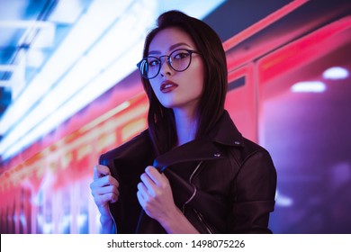 Fashion Beauty Shooting Of Gorgeous Asian Model. Young Beautiful Woman In Black Jacket Posing Over Night City Dramatic Red And Blue Neon Background