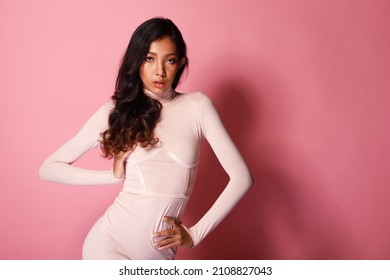 Fashion Beauty Woman Has Long Curl Black Hair Looks At Camera And Express Feeling Smile Happy. Portrait Of Asian Girl Show Tanned Skin Wear Pink Skinny Dress Over Pink Color Block Background