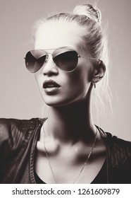 Fashion Black And White Portrait Of A Blonde Woman With Glasses, Black Leather Jacket, Hair Bun. Fashion Sexy And Sensual Look. Gray Background.