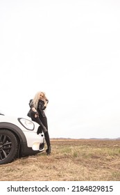 Fashion Blonde Long Curly Hair Lady Sitting On White Automobile Hood Surrounded By Dry Yellow Grass Natural Field. Glamour Rock Woman Posing On Car Transportation Enjoy Freedom Travel Journey Scenery