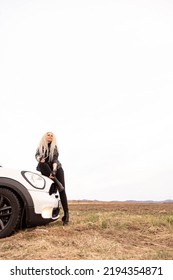 Fashion Blonde Long Curly Hair Lady Sitting On White Automobile Hood Surrounded By Dry Yellow Grass Natural Field. Glamour Rock Woman Posing On Car Transportation Enjoy Freedom Travel Journey Scenery