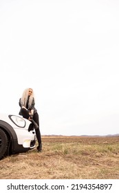 Fashion Blonde Long Curly Hair Lady Sitting On White Automobile Hood Surrounded By Dry Yellow Grass Natural Field. Glamour Rock Woman Posing On Car Transportation Enjoy Freedom Travel Journey Scenery