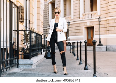 Fashion Blonde Woman In Sunglasses Is Walking On Street On High Heels. She Wears White Jacket And Black Ripped Jeans