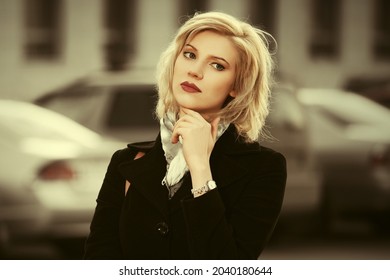 Fashion blonde woman walking on city street Stylish female model in classic black coat and white scarf