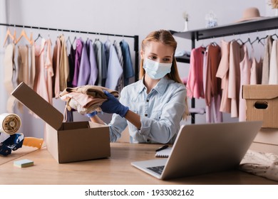 Fashion Boutique Owner In Medical Mask, Packing Clothes Into Boxes Near Laptop On Blurred Foreground