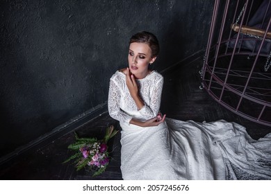 Fashion bride in a white openwork dress without a veil sitting by an iron cage, bars. Unhappy young woman near black textured background. Fictitious marriage. Wedding of convenience. Copy space