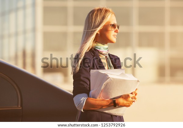 Fashion\
business woman with financial papers next to her car Stylish female\
model wearing dark blue blazer and\
sunglasses