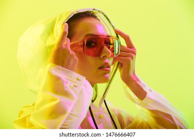 Fashion close up portrait of an attractive young asian woman with long wet hair standing isolated over yellow background, posing in transparent raincoat and sunglasses
