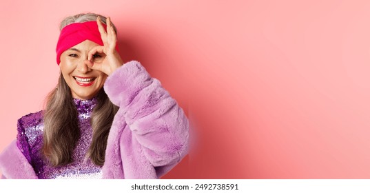 Fashion. Close-up of fashionable asian senior woman smiling, showing OK sign over eye and looking happy at camera, standing over pink background. - Powered by Shutterstock