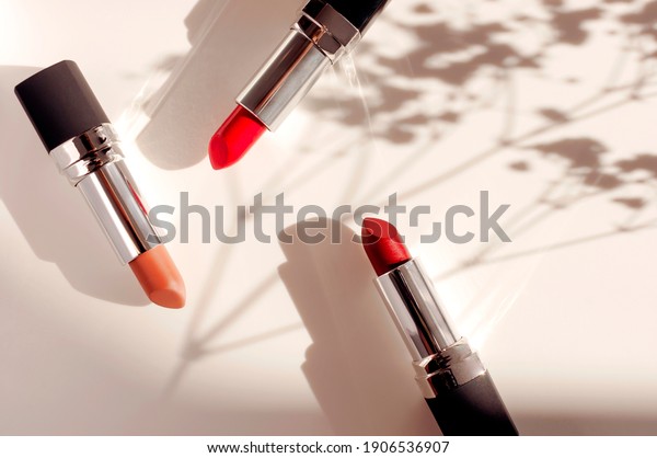 Fashion colorful lipsticks sun shadows from\
flowers on beige background flat lay top view. Beauty and cosmetics\
background. Decorative cosmetics makeup women\'s lipstick beauty\
brand product design