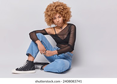 Fashion concept. Studio shot of young pretty fit African american model sitting on floor on white background wearing stylish blue jeans black top mesh turtleneck and sneakers with positive emotions - Powered by Shutterstock