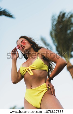 Fashion cute young teenage girl or woman posing and having fun on the beach under palm trees in summer swimsuit . Trendy girl posing. Funny and positive woman in sunglasses