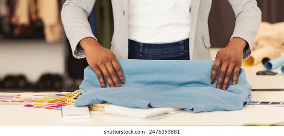 A fashion designer black woman stands at a bright work table, meticulously folding a piece of blue fabric with precision. The workstation is scattered with various sewing accessories, cropped - Powered by Shutterstock