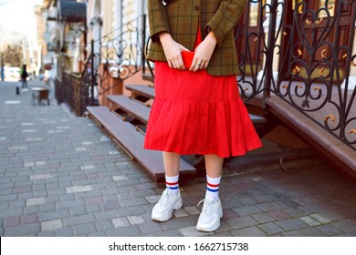 Fashion Details Of Stylish Hipster Blogger Woman Posing On The Street, Wearing White Ugly Sneakers, Long Funny Socks, Red Midi Skirt And Oversized Jacket, Mid Season Time.