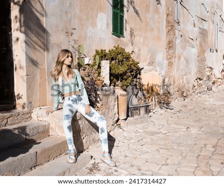 Fashion e-commerce photography: Caucasian blond woman, sitting down in front of a traditional mallorquin finca. Wearing light green top and white pants with blue flowers details 