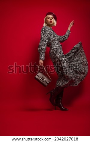 Fashion full length portrait of elegant woman wearing trendy zebra print midi dress, marsala color high leather boots, beret, holding stylish handbag. Model posing in studio, on red background