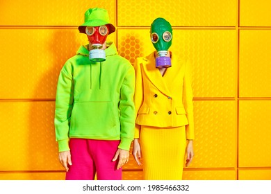 Fashion Of The Future. Portrait Of A Woman And A Man In Bright Clothes And Gas Masks Posing By A Yellow Industrial Wall. Environmental Disaster, Apocalypse.