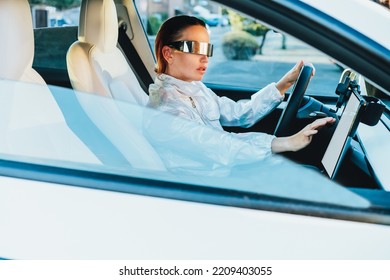 Fashion Girl Driving A White Supercar. Red Hair Woman With Futuristic Eyeglasses Sits By The Car Wheel And Uses An Electronic Dashboard, Tablet Computer Future Technology Concept. Selective Focus.