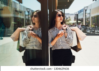 Fashion Girl In Sunglasses Standing Outdoor Holding Cellphone Searching Right Direction Cloth Shop Online Map App On Internet. Young Asian Lady Carrying Shopping Bag In Shopping Mall Center