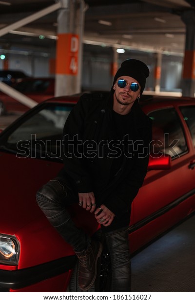 Fashion handsome model man in fashionable clothes\
with a black coat and a hat in stylish sunglasses sits near a red\
car in the city