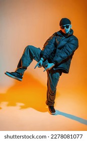 Fashion handsome young hip hop dancer guy with sunglasses and a hat in fashionable clothes with a down jacket and sneakers is dancing in a color studio with orange and neon light - Shutterstock ID 2278081077