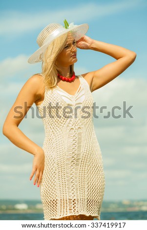 Fashion, happiness and lifestyle concept. Portrait of lovely blonde girl in summer hat wearing white lace dress and red beads on blue sky background outdoors