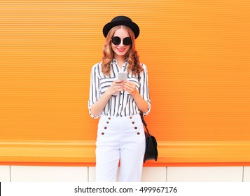 Fashion Happy Young Smiling Woman Model Using Smartphone Wearing A Black Hat White Pants Over Colorful Orange Background