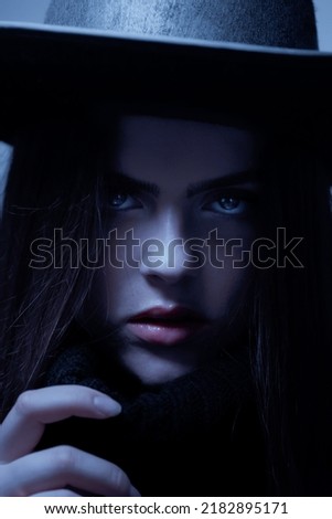 Fashion, horror and make-up concept. Portrait of beautiful woman reaching one hand and wearing black hat in partly face covered with shadow. Blue light filter applied imitation of moonlight