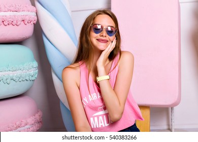 Fashion Indoor Trendy Portrait Of Amazing Beautiful Young Woman Posing Near Big Fake Macaroons And Candy. Happy Blonde Girl With Hearts Sunglasses Holding Hand Near Face. Satisfied Smile.