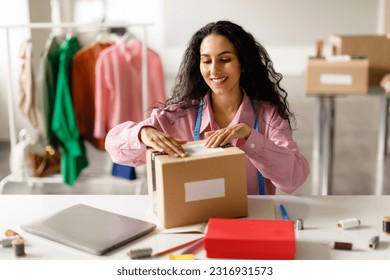 Fashion Industry Entrepreneurship. Happy Middle Eastern Woman Packing Designer's Clothes In Cardboard Box Sitting At Desk In Fashionable Store Indoor. Clothing Boutique Ownership - Powered by Shutterstock