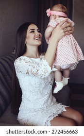 Fashion Interior Photo Of Beautiful Mother With Long Dark Hair In Elegant Lace Dress Holding Her Little Cute Baby Girl In Hands