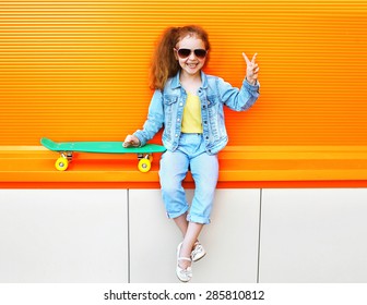 Fashion kid. Stylish little girl child wearing a summer or autumn jeans clothes, sunglasses with skateboard having fun in city over colorful orange background