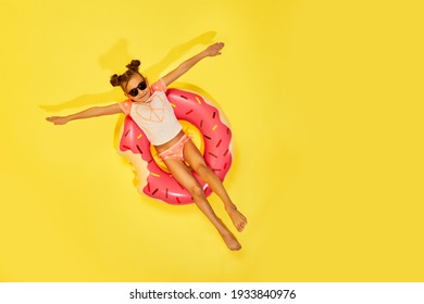 Fashion Little Child Girl In Swimwear Lying On Pink Donut Rubber Ring On Yellow Background. Top View. Copy Space