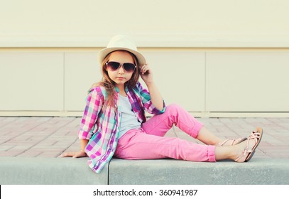 Fashion little girl model wearing a pink checkered shirt, hat and sunglasses in city