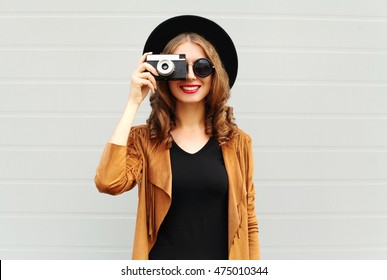 Fashion look, pretty cool young woman model with retro film camera wearing a elegant hat, brown jacket, curly hair outdoors over city grey background