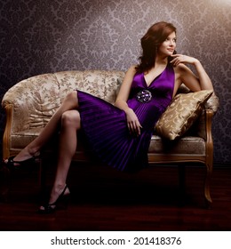 Fashion Luxury Model In Purple Dress. Young Beauty Style Girl. Beautiful Luxurious Woman Sitting On A Gold Vintage Couch 