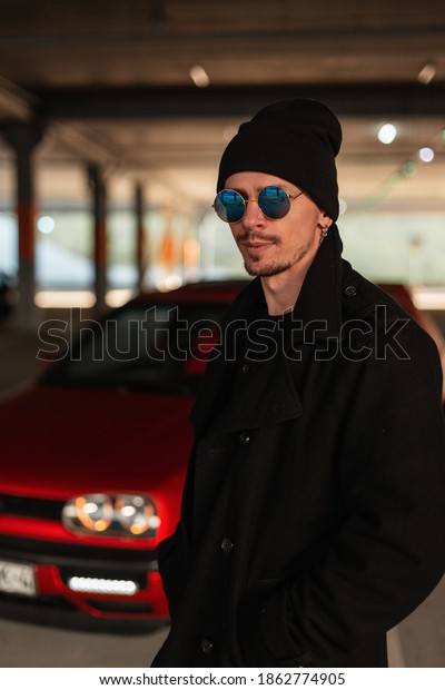 Fashion man with sunglasses in black stylish\
coat and hat near a red car on the\
street