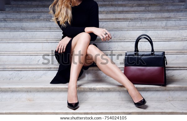 Fashion model in black\
dress with big leather red handbag in high heel shoes sitting on\
the white stair