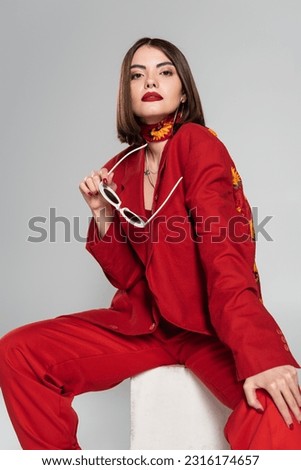 fashion model with brunette short hair and nose piercing posing in red suit while holding sunglasses and sitting on concrete cube on grey background, stylish posing, lady in red, young woman