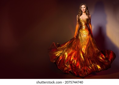 Fashion Model in Long Golden Dress. Smiling Beauty Woman in Evening Shining Fluttering Gold Gown walking over Dark Brown Background