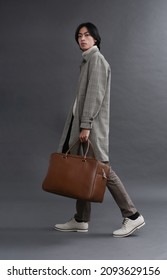 Fashion Model. Man With Hairstyle In Coat Holding Handbag Walking In Studio On Gray Background

