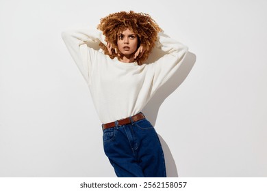 Moda, modelo, retrato, estudio, estilo casual, belleza Una mujer joven con el pelo rizado posa en un suéter blanco y los pantalones vaqueros azules contra un fondo blanco liso, exhibiendo la ropa casual moderna