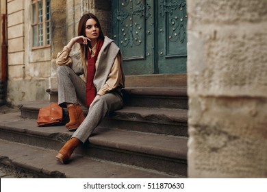 Fashion Model In Street. Beautiful Sexy Woman In Stylish Fashionable Fall Clothes: Shirt, Scarf, Pants, Sweater, Shoes Sitting On Stairs In Spring. Girl In High Fashion Autumn Clothing Posing Outdoors